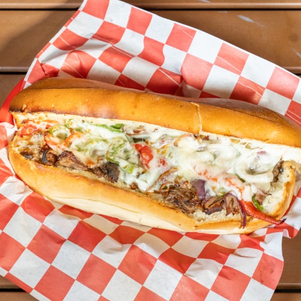 A Cali-Forno Pizzeria cheesesteak sandwich with melted cheese, peppers, and onions on a red checkered paper.