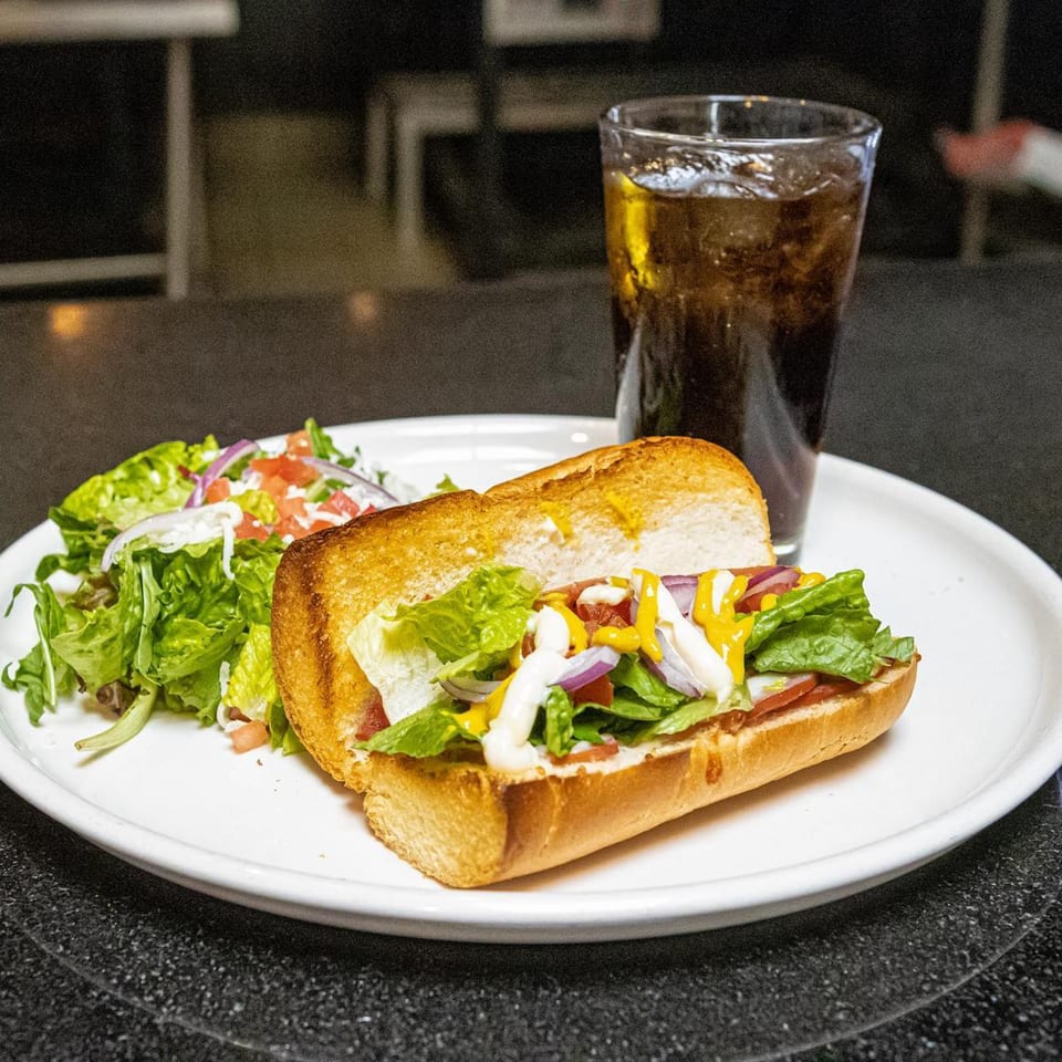 A Cali-Forno Pizzeria sandwich with salad and a drink on a white plate.