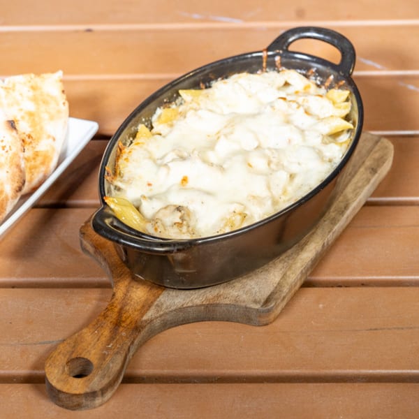 Baked pasta with melted cheese in a black oval dish on a wooden board, with flatbread on the side.